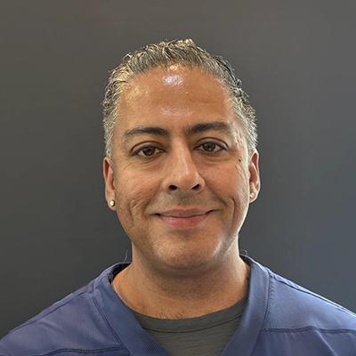 Headshot of Vic Sarda wearing blue medical scrubs, facing the camera against a plain dark background.