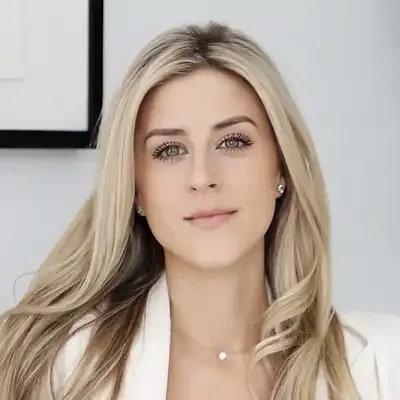 Professional headshot of a person with long light-colored hair, wearing a white blazer and a delicate necklace, photographed against a clean, light-colored indoor background.